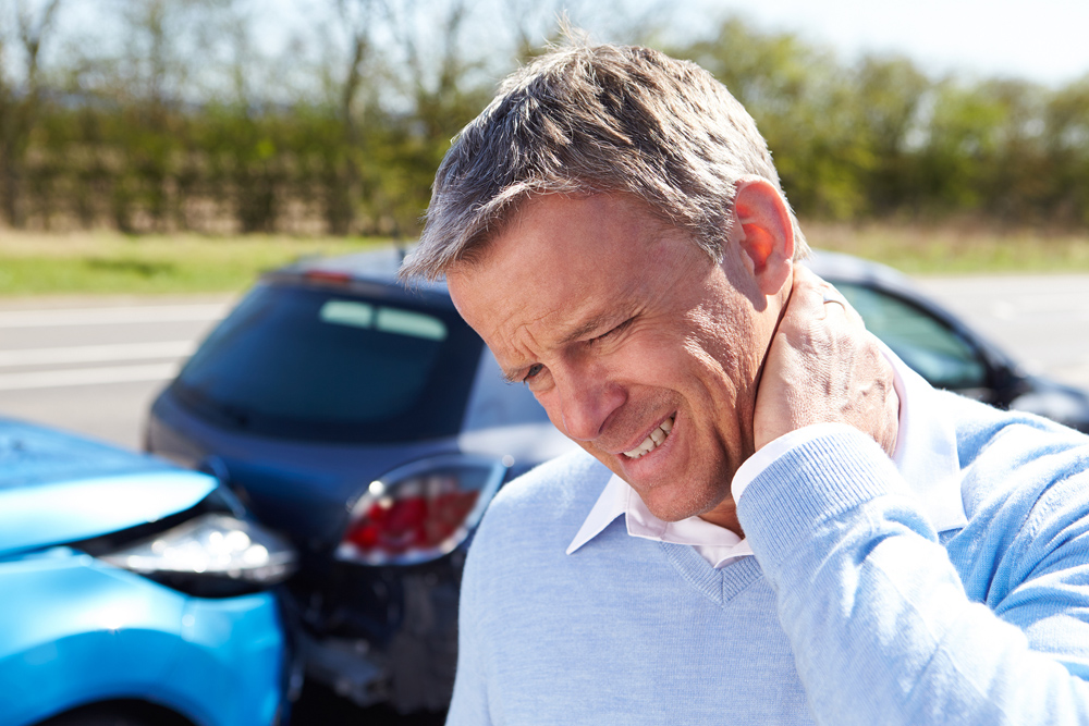 man holding his neck becuase of whiplash pain with a car accident in the background